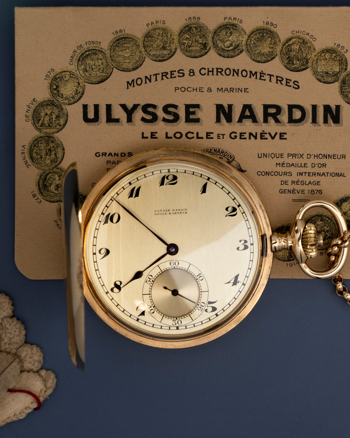 An open gold-toned Ulysse Nardin pocket watch with a cream dial, placed in front of branded packaging on a dark blue background.