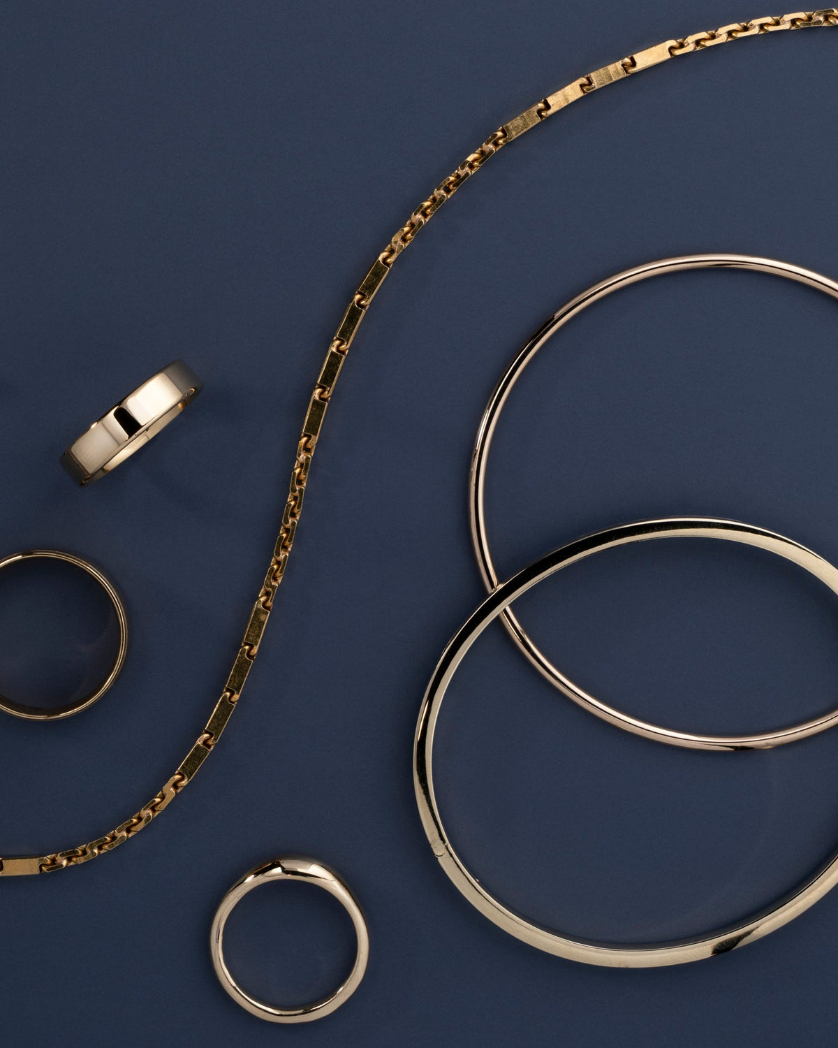 A styled flat-lay of gold-toned jewelry on a dark navy blue background, featuring a chain necklace, two bangles, and three rings.